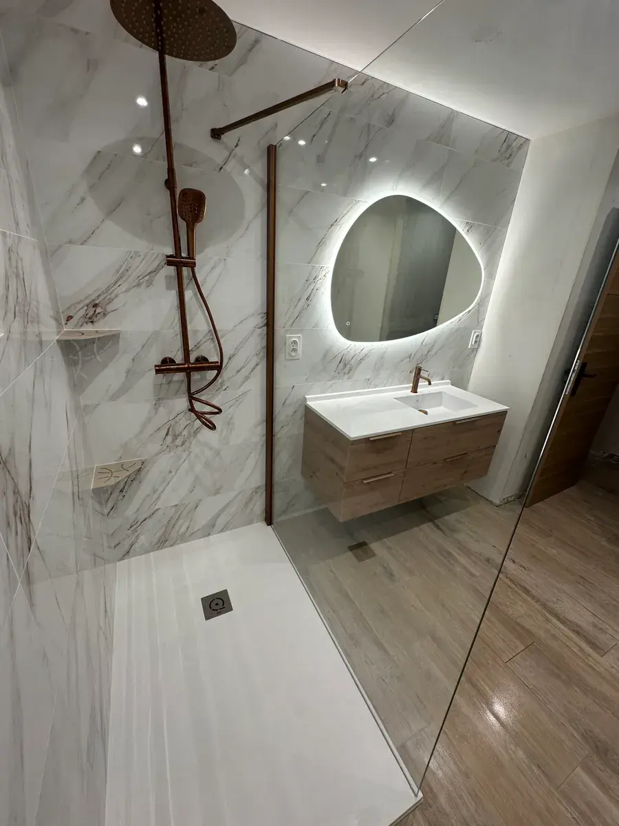 Douche à l'italienne avec colonne de douche cuivrée, faïence effet marbre et miroir galet rétroéclairé au-dessus d'un meuble vasque en bois